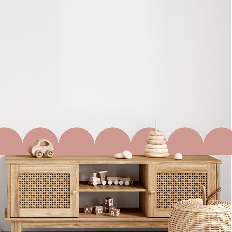 May include: A light wood cabinet with woven doors and open shelves displays wooden toys, including a car, train, and stacking rings. The cabinet is set against a white wall with a pink scallop design.