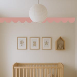 May include: A nursery interior with a light wood cot, framed art prints, and a decorative scalloped border in a soft pink colour. A paper lantern hangs from the ceiling. The art includes a hot air balloon, a teddy bear, and a star.