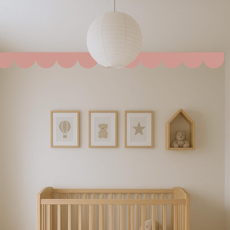 May include: A nursery interior with a light wood cot, framed art prints, and a decorative scalloped border in a soft pink colour. A paper lantern hangs from the ceiling. The art includes a hot air balloon, a teddy bear, and a star.