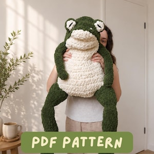 Può includere: Un grande peluche a forma di rana lavorato all'uncinetto, di colore verde scuro e crema, con arti lunghi e occhi prominenti. La rana ha un aspetto testurizzato e soffice. Un riquadro grafico verde in basso riporta il testo PDF PATTERN.