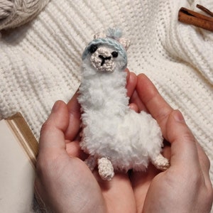 May include: A white crocheted llama wearing a light blue crocheted hat with a white pom-pom. The llama is being held in the palms of two hands.