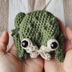 May include: A small, handmade crochet frog plush toy featuring a textured olive green body, two large black plastic safety eyes, and light beige yarn detailing around the eyes and mouth area. The stuffed creature has a rounded shape with small limbs.