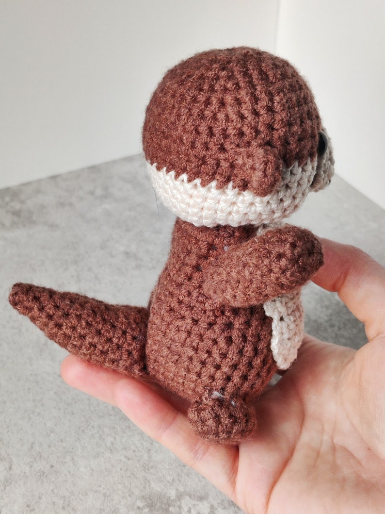 May include: A brown crocheted otter soft toy with white accents on the face and chest. The otter is standing on its hind legs with its tail curled up.