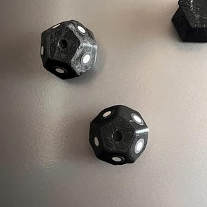 May include: Black, multi-sided dice with white dot markings. The dice have a textured surface and a hole in the center of one side. The dice are likely used for tabletop games or role-playing games.