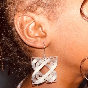 Puede incluir: Primer plano de una oreja con un pendiente cuadrado blanco. El pendiente presenta un diseño geométrico de líneas entrelazadas y un gancho plateado. El pendiente es el foco principal de la imagen.