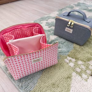 Pode incluir: Duas nécessaires de maquilhagem, uma rosa com um padrão de bolinhas brancas e outra azul com uma textura tecida. Ambas as nécessaires têm fechos de correr e alças douradas.