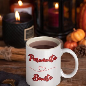May include: White ceramic mug with a red heart and the text "Permanita Bonita" in red cursive lettering. The mug is filled with a brown liquid and is sitting on a gray napkin.