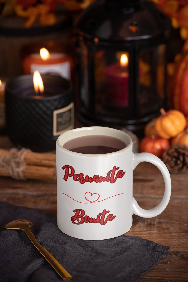 May include: White ceramic mug with a red heart and the text "Permanita Bonita" in red cursive lettering. The mug is filled with a brown liquid and is sitting on a gray napkin.