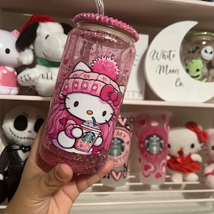 Puede incluir: Vaso de vidrio rosa lleno de purpurina con un diseño de Hello Kitty, sosteniendo una taza de Starbucks. El vaso tiene una tapa rosa con un borde de pedrería y una pajita a juego. Otros artículos con temática de Hello Kitty son visibles en el fondo.