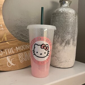 Puede incluir: Un vaso Hello Kitty rosa y blanco con purpurina. El vaso tiene una tapa transparente y una pajita rosa. El vaso tiene un gráfico de Hello Kitty con el texto "Hello Kitty" en él.