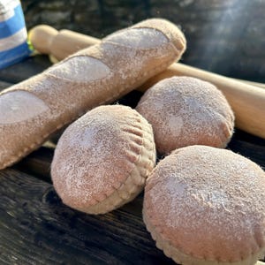 Puede incluir: Una colección de productos de panadería de felpa, que incluye una baguette y tres panecillos redondos, espolvoreados con polvo blanco. Un rodillo de madera descansa detrás del pan. Los artículos están dispuestos sobre una superficie de madera oscura, lo que sugiere un tema de panadería o cocina.
