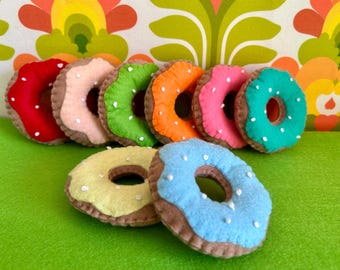 May include: A collection of colorful felt donuts with white sprinkles. The donuts are in various colors, including red, pink, green, orange, yellow, and blue, with brown edges. They are arranged on a green surface.