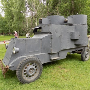 May include: A grey armoured car with a large, boxy body and a chain hanging from the front. The car has a large, open top and a small, open door on the side. The car is sitting on a grassy field.