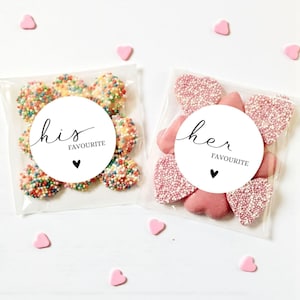 May include: Two cellophane bags of sweets, one with colourful sprinkles and the other with pink heart-shaped candies. Each bag has a white circular label with the words "his" and "her" and the word "FAVOURITE" below, with a small black heart. Pink heart-shaped sweets are scattered around.