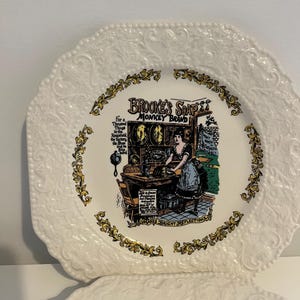 Peut inclure: Assiette octogonale blanche avec un motif floral en relief et une illustration centrale. L'illustration présente le texte "Brooke's Soap Monkey Brand" et une femme travaillant sur une machine. Des accents floraux dorés entourent l'illustration.