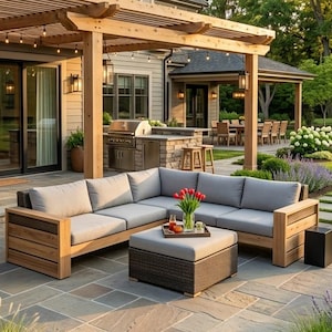 May include: A wooden sectional sofa with light gray cushions and a matching square ottoman on a stone patio. The furniture is positioned under a wooden pergola attached to a house, featuring a built-in outdoor kitchen and garden landscaping.