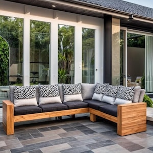 May include: A modern outdoor patio with a large sectional sofa made of wood and black cushions. The sofa is positioned in front of a large window and a wooden wall with a circular cutout. There are black metal chairs and a table with a glass top in the background.