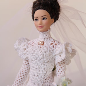 Puede incluir: Muñeca con un vestido de novia blanco de ganchillo con cuello alto y detalle de perlas. La muñeca tiene el pelo oscuro recogido en un moño y un velo transparente. Sostiene un ramo de flores blancas.