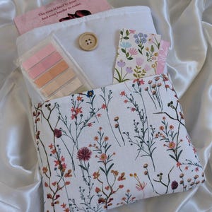 May include: A white fabric pouch with a floral print featuring small pink, red, and yellow flowers. The pouch has a button closure and a pocket on the front. The pouch is filled with a pink book, sticky notes, and a card with a floral print.
