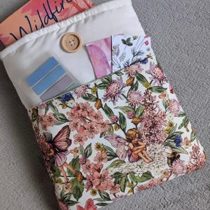 May include: A white fabric book sleeve with a floral pattern featuring fairies and flowers. The sleeve has a pocket on the front with a wooden button, blue sticky notes, and a pink and white patterned card.