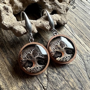 Può includere: Un paio di orecchini rotondi in legno con un design ad albero della vita. Gli orecchini hanno ganci color argento e una finitura trasparente e lucida sull'immagine dell'albero. Il design dell'albero è nei toni del marrone e beige.