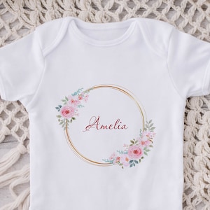 May include: White baby onesie with a gold circle and pink floral design. The name Amelia is written in burgundy inside the circle.