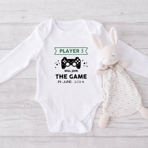 May include: White long-sleeved baby onesie with a green banner that says "Player 3" and a black video game controller graphic. The text below the controller says "Will join the game in June, 2024".