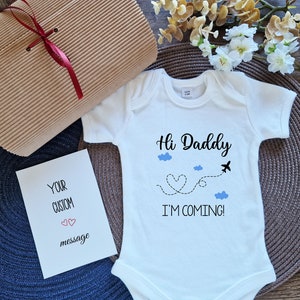 May include: White baby onesie with the text "Hi Daddy I'M COMING!" and a graphic of a plane and clouds. Includes a gift box and a card that says "YOUR CUSTOM message".