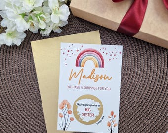 May include: A greeting card with the name "Madison" and the text "WE HAVE A SURPRISE FOR YOU" is displayed. The card has a rainbow illustration and the words "You're going to be a BIG SISTER". A gold envelope and a gift box with a burgundy ribbon are also visible.