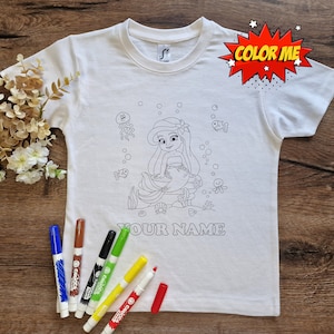 Peut inclure: T-shirt blanc avec un contour noir et blanc d'une sirène avec le texte "Color Me" et "Your Name" imprimé sur le devant. Le t-shirt est accompagné de plusieurs marqueurs colorés.