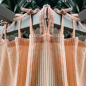 May include: Close-up of several orange and white striped curtains hanging from a gray wooden structure. The curtains are tied at the top with matching orange and white striped ties. The background is blurred green foliage.