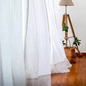 Puede incluir: Cortinas de lino blanco con una textura sutil cuelgan en una habitación con suelo de madera. Las cortinas están reunidas en la parte superior y cuelgan holgadamente, creando una sensación suave y aireada.