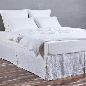 Może przedstawiać: Biała pościelowa spódnica z lnu z falbanką i wiązaniami w rogach. Spódnica jest na białym łóżku z białą pościelą i poduszkami z lnu.