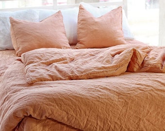 May include: A bed with peach-colored linen bedding, including a duvet and two pillows. The pillows are peach-colored and white. The bedding is arranged on the bed in a casual, inviting manner, with soft folds and wrinkles.