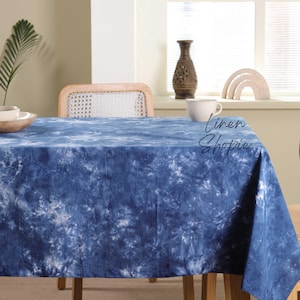 May include: A blue and white tie-dye tablecloth on a wooden table with a white wicker chair. The tablecloth has a light blue background with white tie-dye patterns. The text "Linen Shopie" is visible in the image.