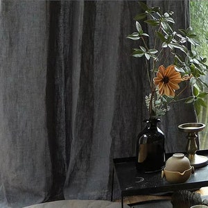 May include: A black vase with a single orange flower and green leaves sits on a black table with a gold candle holder and a small brown ceramic pot. The table is in front of a window with a view of green trees.