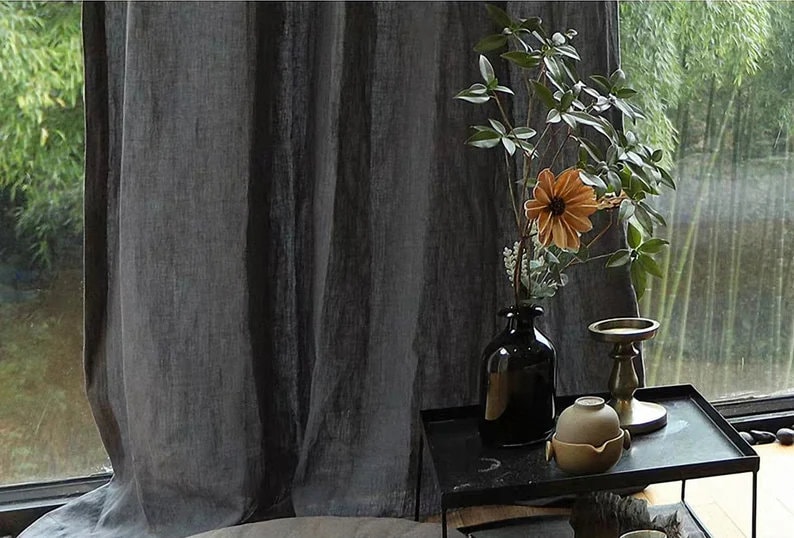 May include: A black vase with a single orange flower and green leaves sits on a black table with a gold candle holder and a small brown ceramic pot. The table is in front of a window with a view of green trees.
