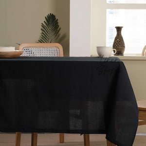 May include: A black linen tablecloth draped over a wooden table with a white cup and saucer on top. The tablecloth is wrinkle-free and has a smooth texture.