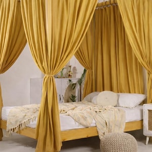 May include: A four-poster bed with a light wood frame and mustard-yellow curtains. The bed has white linens, a patterned throw blanket, and decorative pillows. A woven pouffe sits at the foot of the bed.