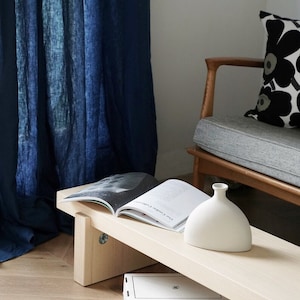 May include: A light wood bench with a white ceramic vase and a book titled "The Cookie Collection" on top. The bench is in front of a blue linen curtain and a wooden chair with a gray cushion and a black and white pillow with a floral pattern.