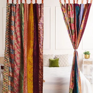 K&ouml;nnte beinhalten: Ein Paar bunte Patchwork-Vorh&auml;nge im Boho-Stil. Die Vorh&auml;nge bestehen aus verschiedenen Stoffen und Mustern und sind an einer schwarzen Gardinenstange aufgeh&auml;ngt. Die Vorh&auml;nge sind in der Mitte zur&uuml;ckgebunden und h&auml;ngen vor einer wei&szlig;en Wand. Im Hintergrund ist ein Bett mit wei&szlig;en Bettw&auml;sche und einem gr&uuml;nen Kissen zu sehen.