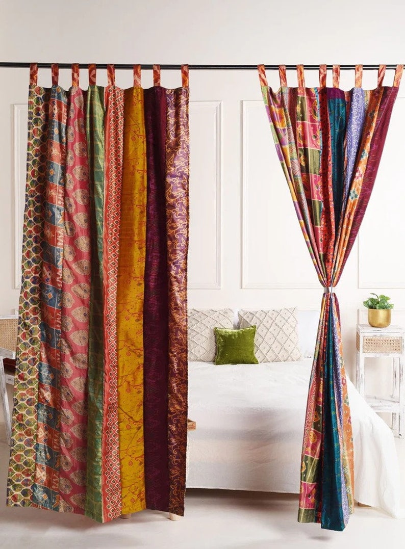 K&ouml;nnte beinhalten: Ein Paar bunte Patchwork-Vorh&auml;nge im Boho-Stil. Die Vorh&auml;nge bestehen aus verschiedenen Stoffen und Mustern und sind an einer schwarzen Gardinenstange aufgeh&auml;ngt. Die Vorh&auml;nge sind in der Mitte zur&uuml;ckgebunden und h&auml;ngen vor einer wei&szlig;en Wand. Im Hintergrund ist ein Bett mit wei&szlig;en Bettw&auml;sche und einem gr&uuml;nen Kissen zu sehen.