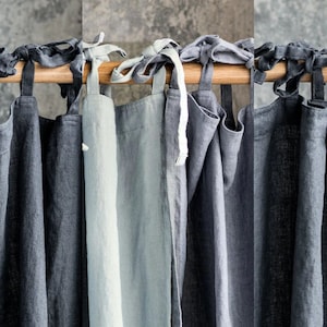 Può includere: Un set di otto tende in lino in varie tonalità di grigio e blu, appese a un'asta di legno. Le tende hanno un design semplice e rustico e sono legate con anelli di tessuto.