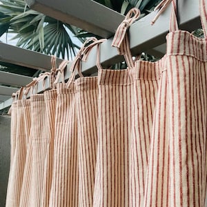 May include: Set of six cream-coloured curtains with thin red stripes. The curtains are hung on a wooden rod and have a simple tie-top design.