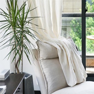 May include: A white linen throw draped over a beige cushion on a chair. A green plant is in the background.