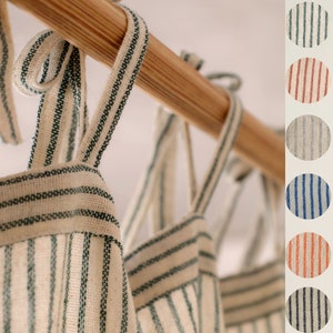 May include: Close-up of several off-white fabric panels with green stripes, hanging from a wooden rod. The panels have tie closures. Color swatches show green, maroon, grey, blue, orange, and black stripe options.