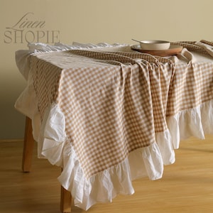 May include: A tan and white gingham tablecloth with a white ruffled edge. The tablecloth is draped over a wooden table, with a bowl and plate visible on top. The text "Linen SHOPiE" is in the upper left corner.