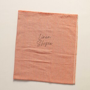 K&ouml;nnte beinhalten: Ein St&uuml;ck orange-wei&szlig; gestreiftes Stoff mit dem Text "Linen Shoppe" darauf.