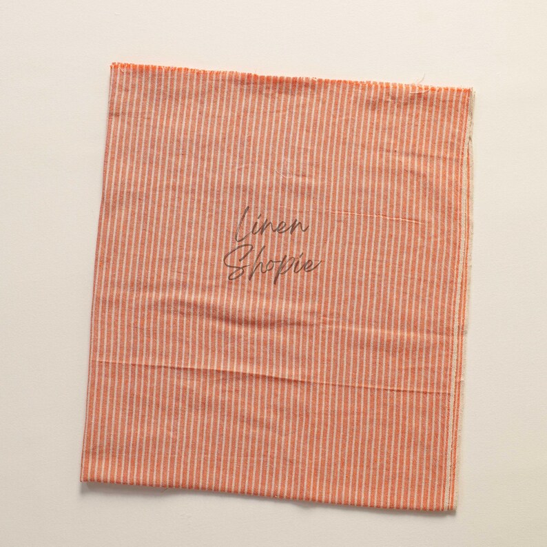 K&ouml;nnte beinhalten: Ein St&uuml;ck orange-wei&szlig; gestreiftes Stoff mit dem Text "Linen Shoppe" darauf.