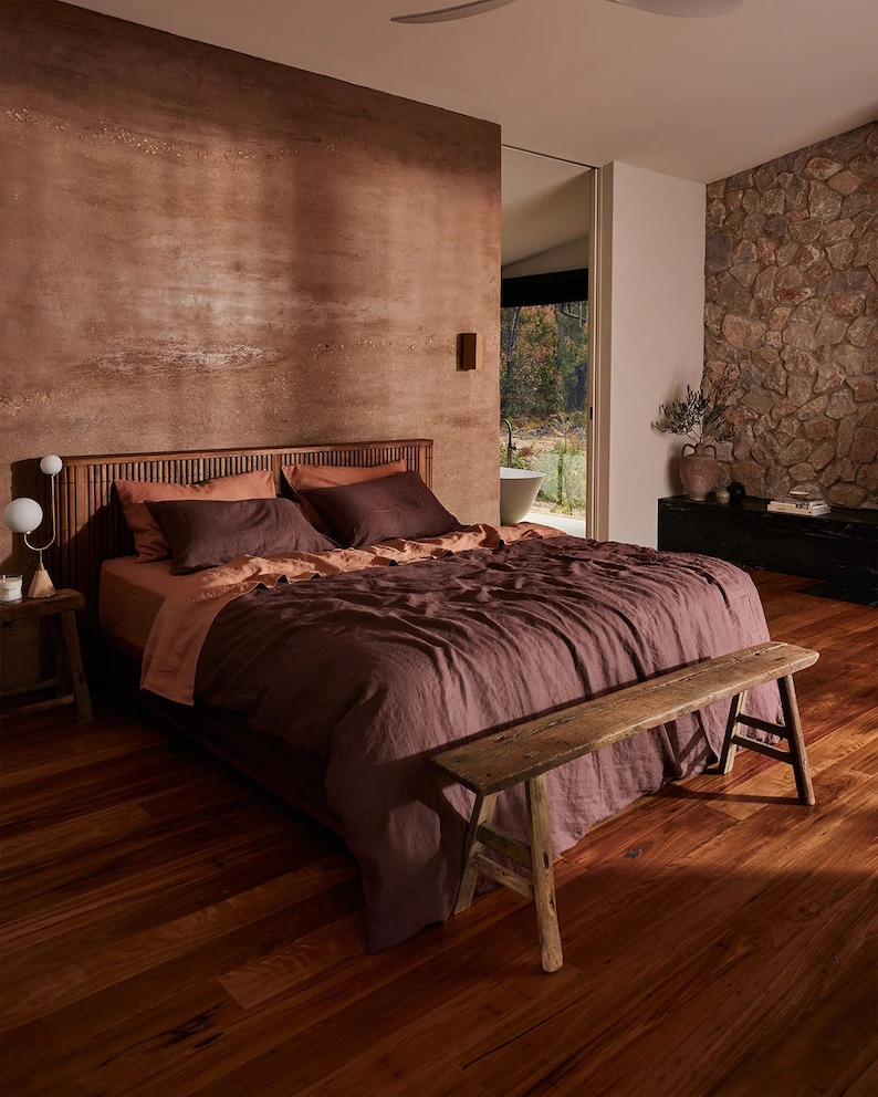May include: A bedroom with a king-size bed featuring a dark brown linen duvet and a wooden headboard. The bed is next to a rustic wooden bench. The walls are a warm brown colour and there is a stone wall in the background.